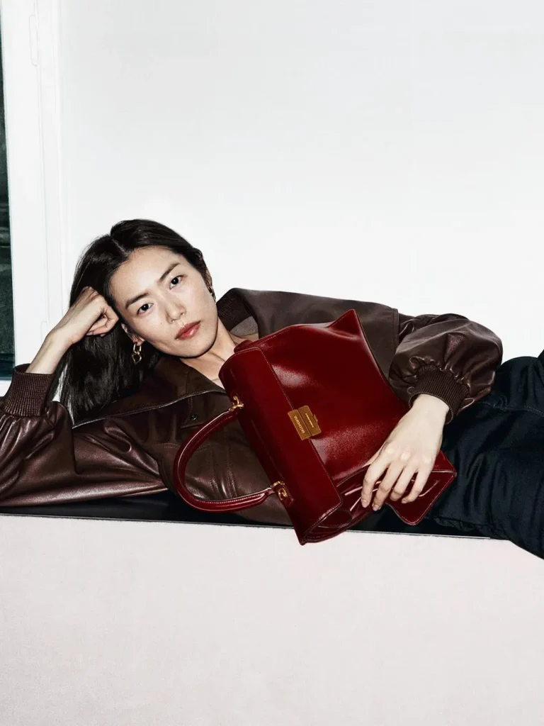 Woman reclining in a brown leather jacket, holding a structured burgundy handbag against a white background