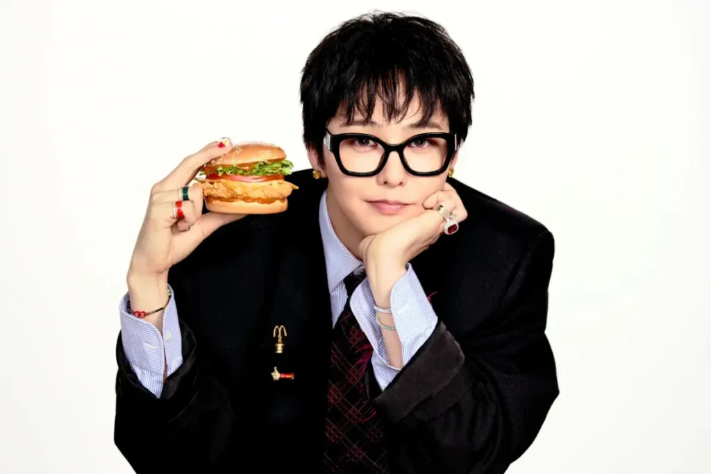 Person in black suit and glasses holds a burger beside their face, resting their chin on one hand against a white background