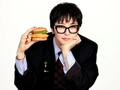 Person in black suit and glasses holds a burger beside their face, resting their chin on one hand against a white background
