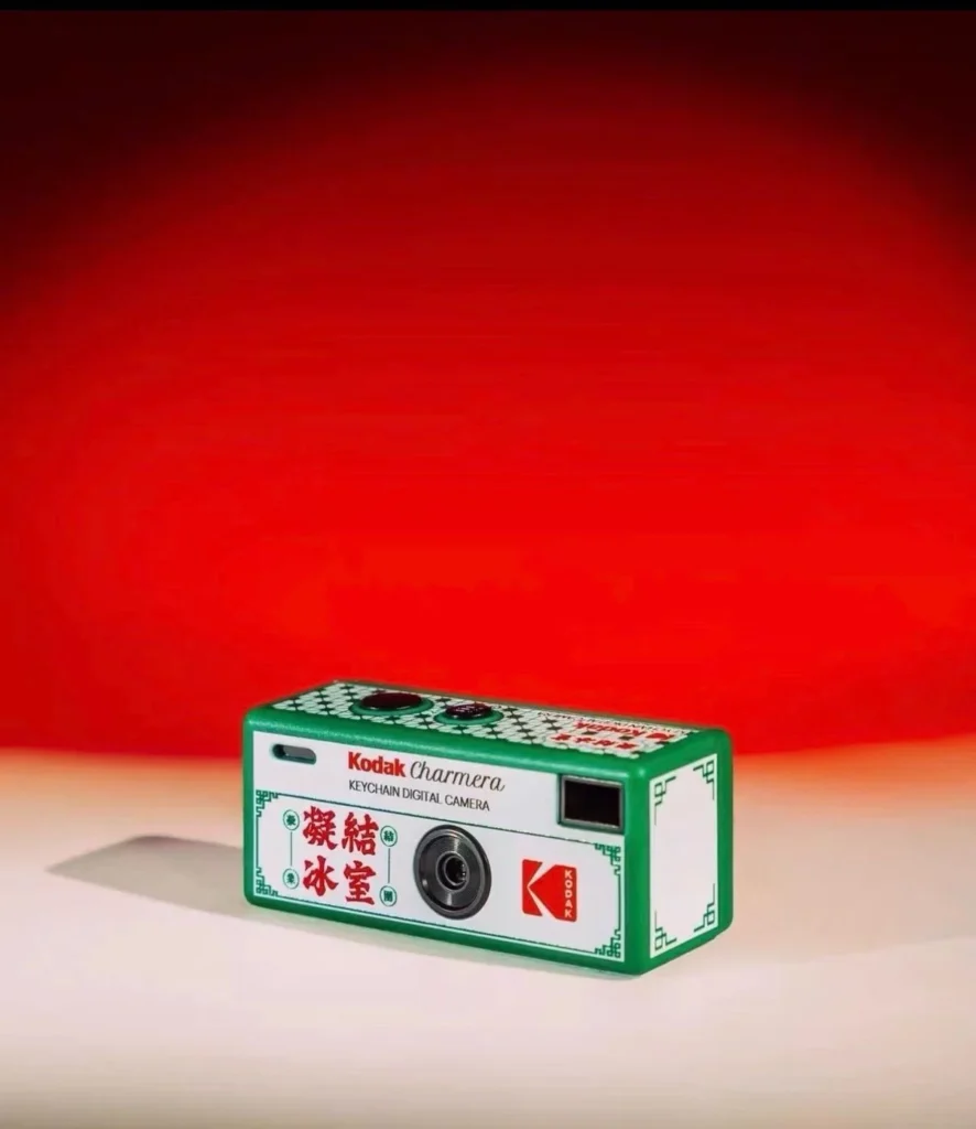 Green-and-white Kodak Charmera keychain digital camera with Chinese characters, on a pale surface against a red backdrop