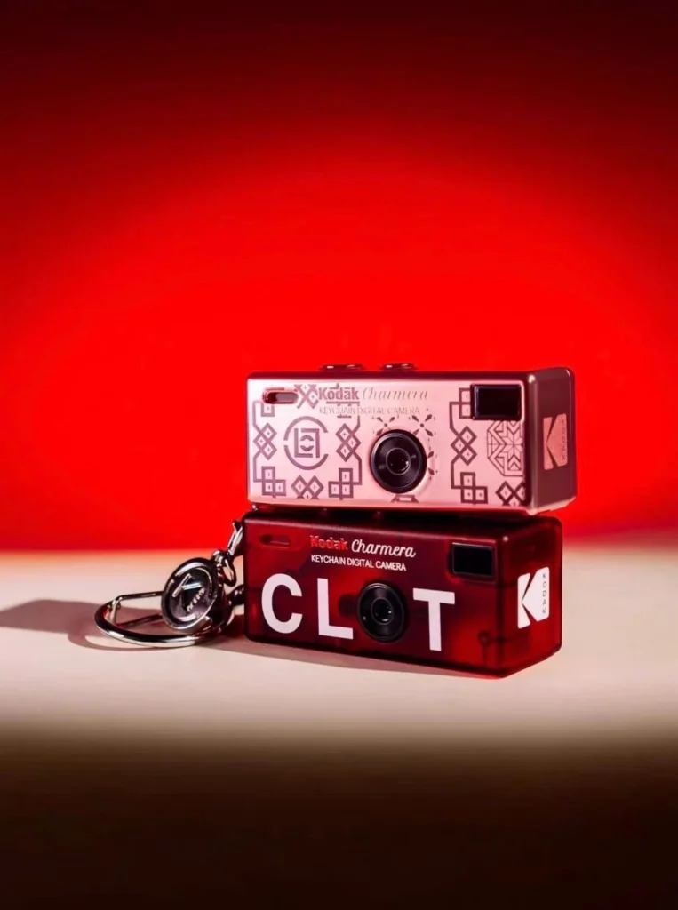Two Kodak Charmera keychain digital cameras, one pink patterned and one red marked CLT, stacked on a tabletop against red