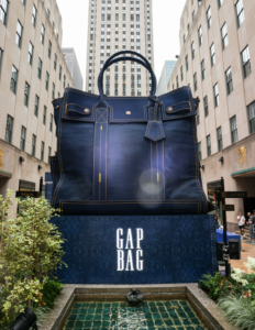 Oversized navy Gap bag with gold stitching displayed above a fountain in a city courtyard flanked by tall buildings