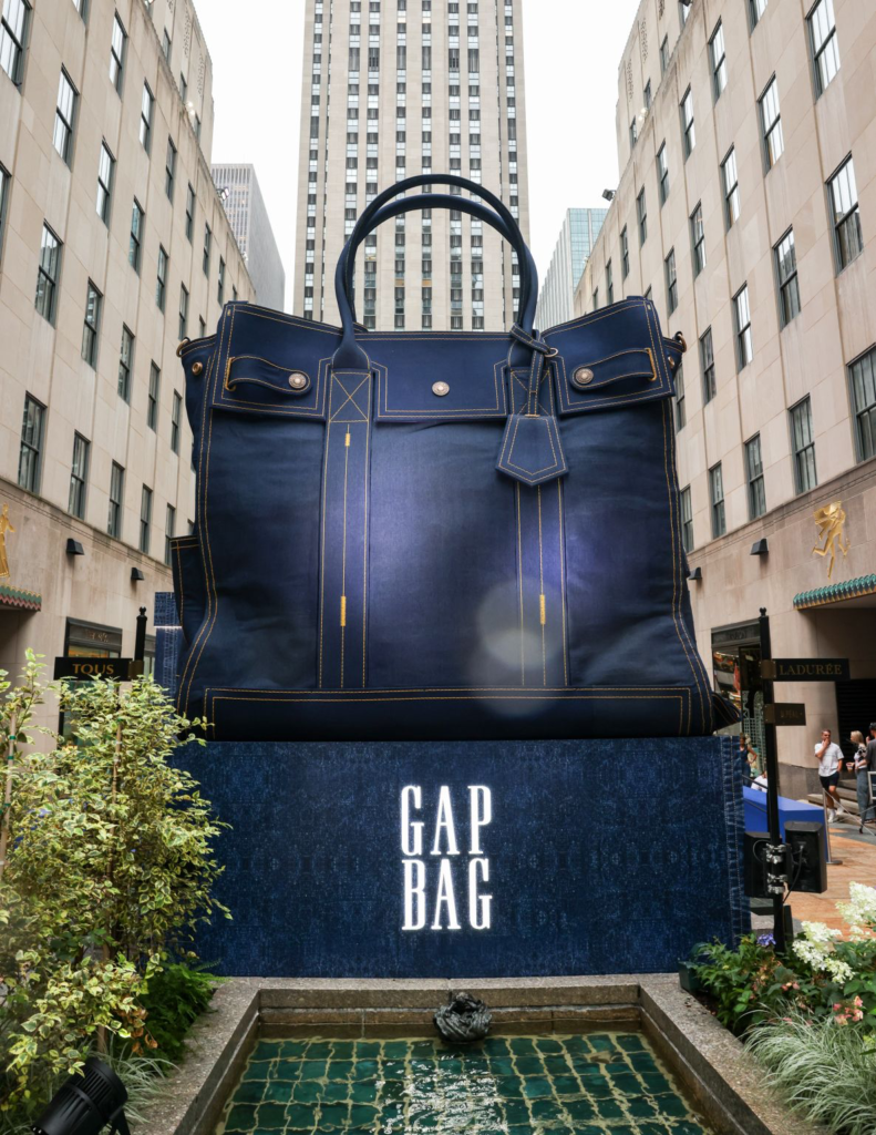 Oversized navy Gap bag with gold stitching displayed above a fountain in a city courtyard flanked by tall buildings