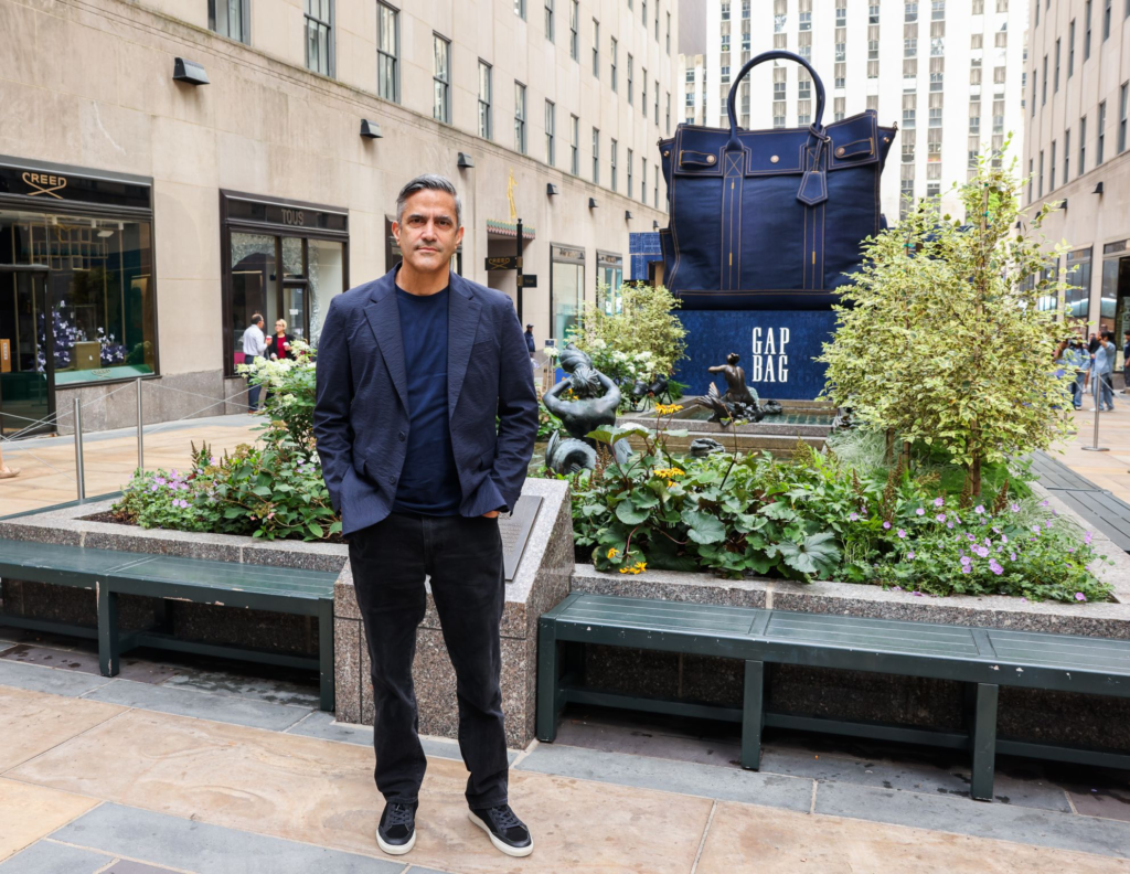 Man in navy clothing stands before a giant navy GAP handbag display in a landscaped Rockefeller Center plaza
