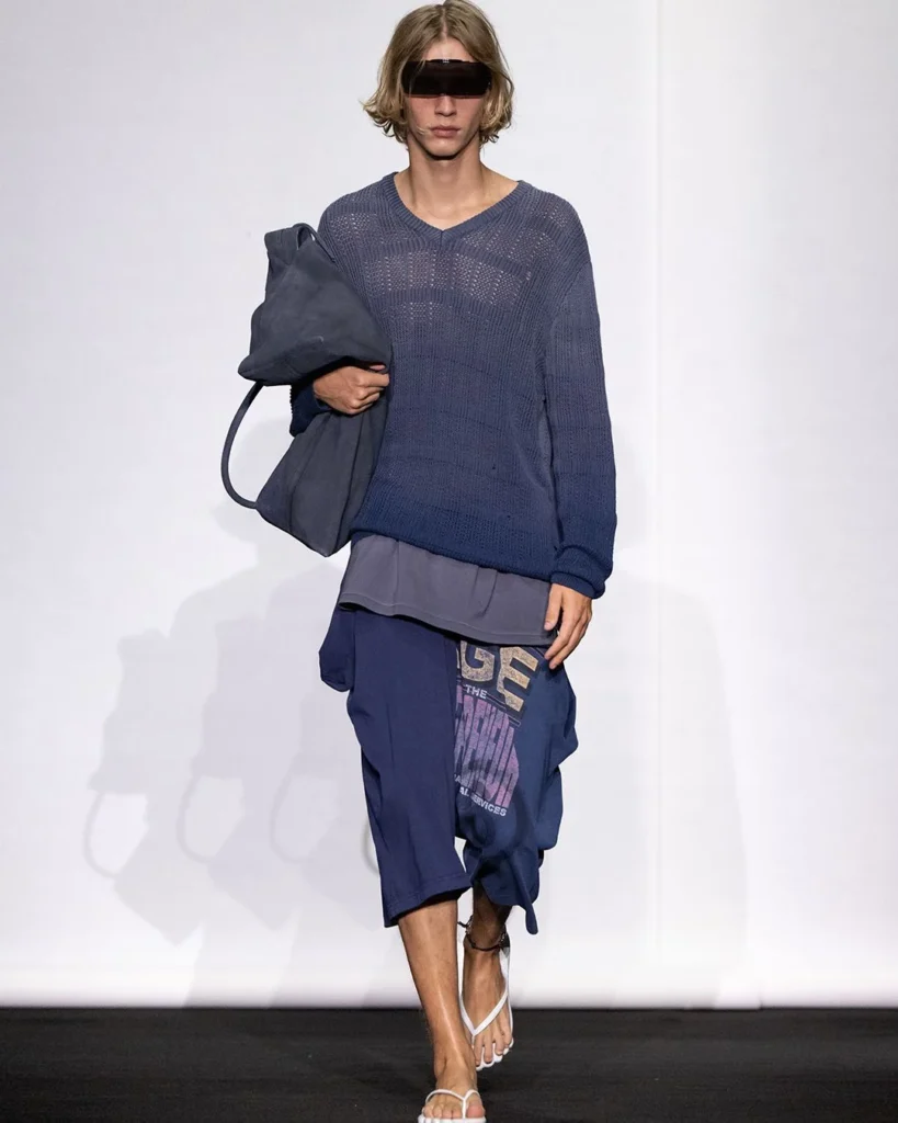 Model in a sheer navy knit sweater, long shorts, white sandals, black visor, and oversized navy bag on a runway
