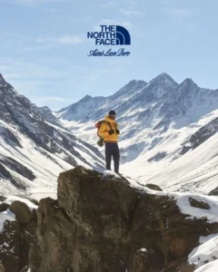 Person in a yellow jacket stands on a rocky outcrop in a snow-covered mountain valley beneath brand logos