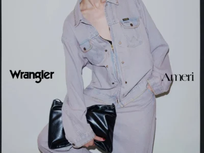 Model in sunglasses wears a pale lavender denim jacket and pants, holding a black leather clutch against a gray backdrop