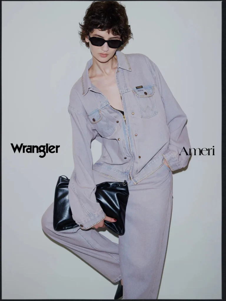 Model in sunglasses wears a pale lavender denim jacket and pants, holding a black leather clutch against a gray backdrop