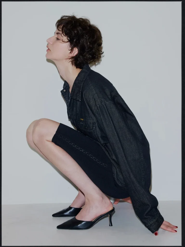 Person crouching in a black denim jacket and skirt with pointed mule heels against a plain gray wall