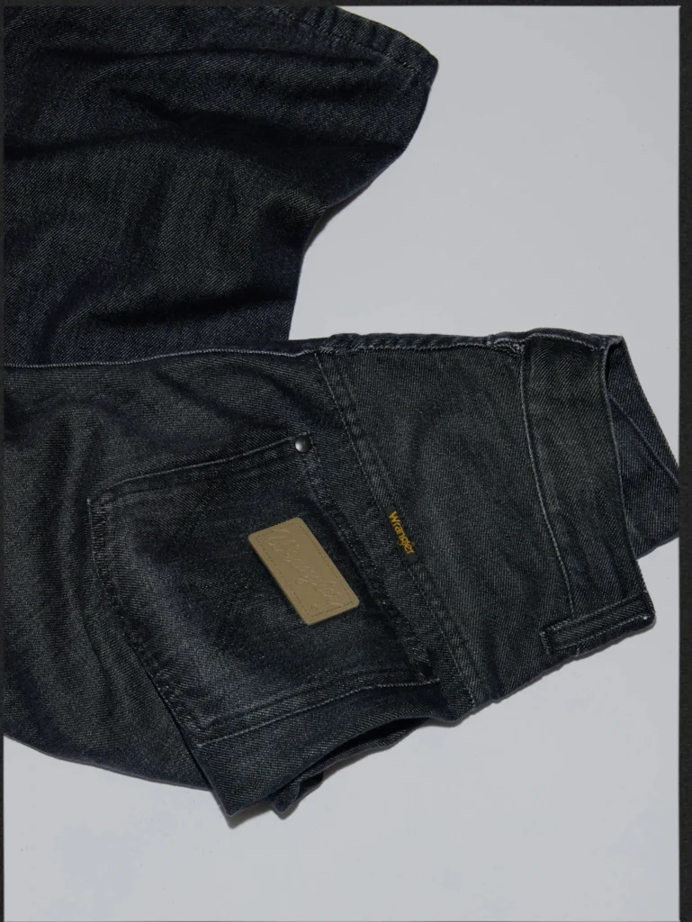 Close-up of folded black denim jeans with a stitched back pocket, metal rivet, and tan leather brand patch on a white surface