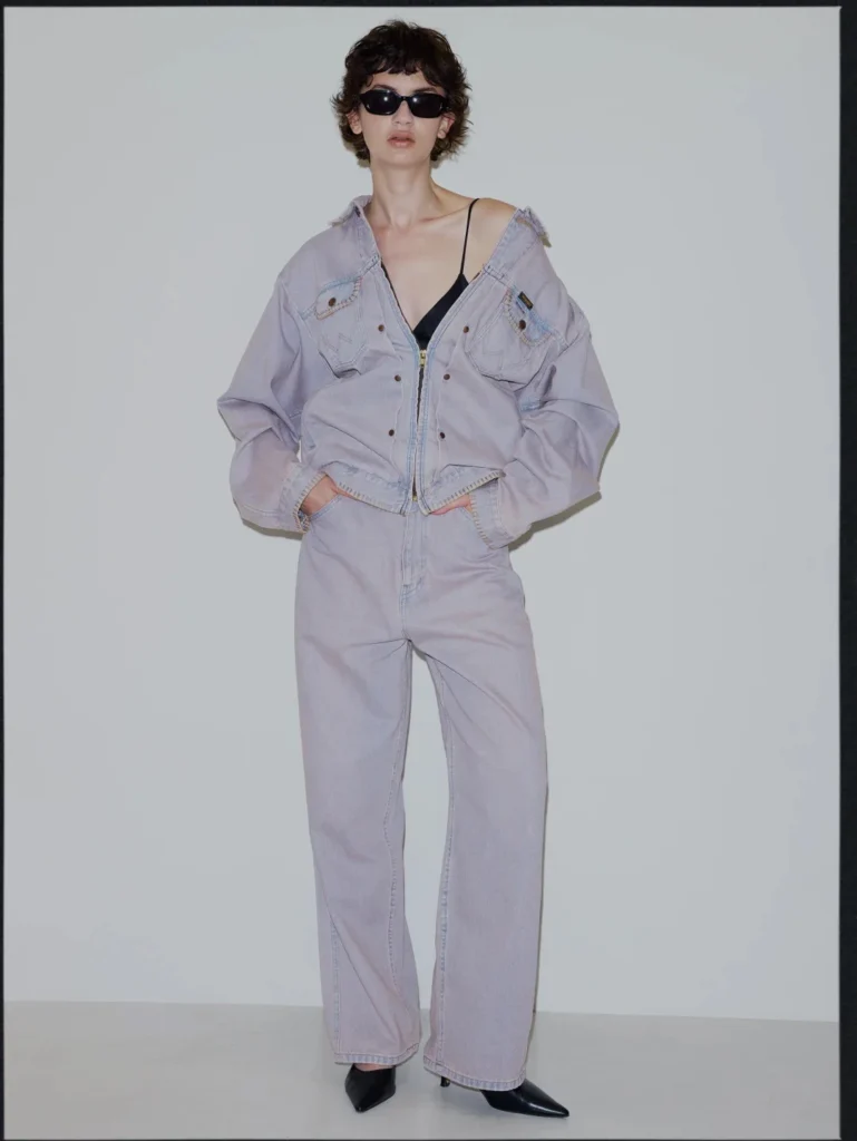 Model wearing a pale lavender denim jacket and wide-leg jeans with black sunglasses and pointed shoes against a gray backdrop