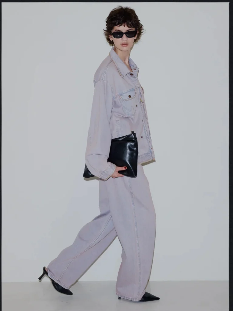 Model in black sunglasses wears a pale denim jacket and wide-leg pants, carrying a black leather clutch against a gray