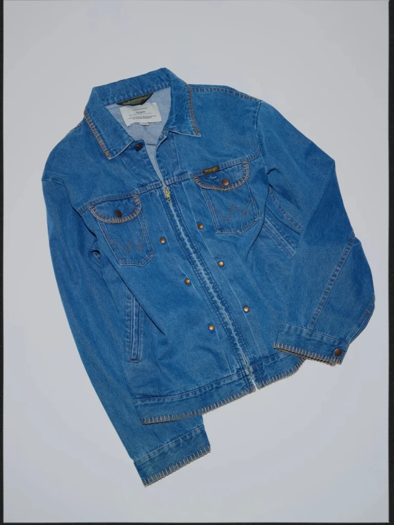 Blue denim jacket with contrasting stitching, flap chest pockets, zip front, and button closures, laid flat on a gray