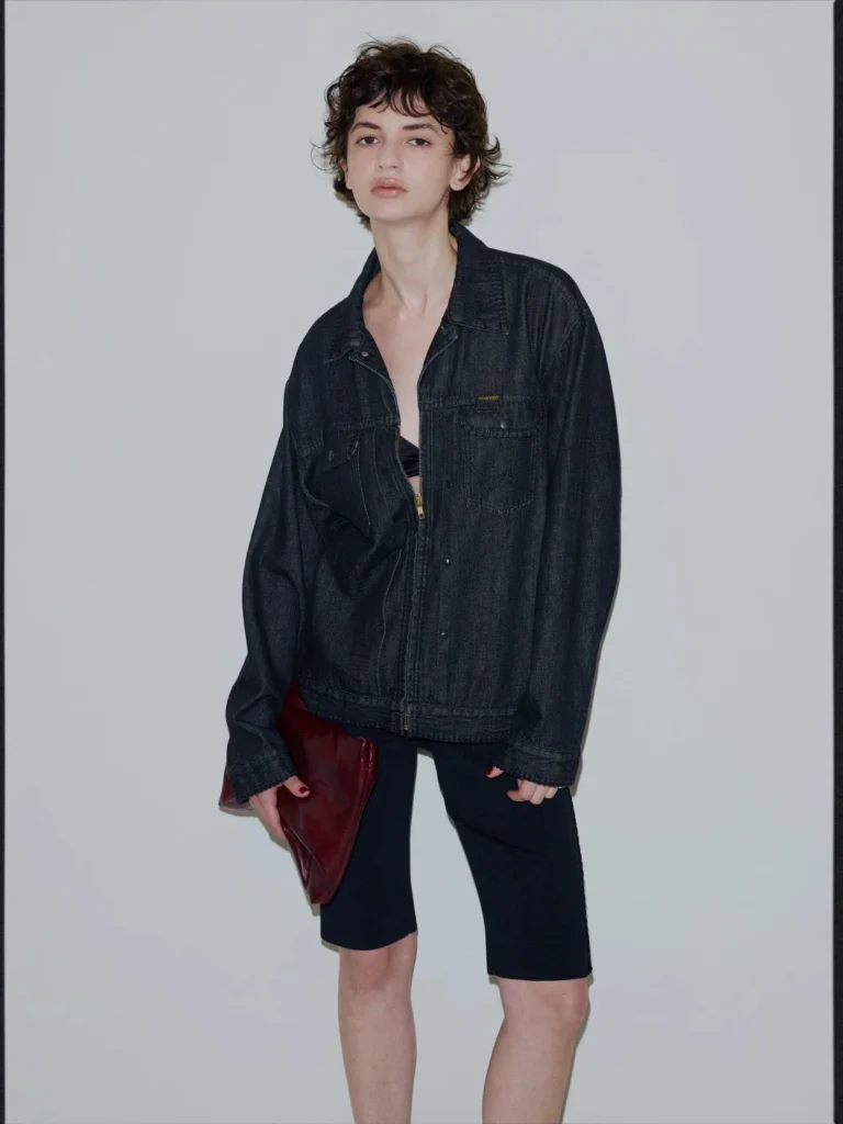 Model wearing a dark denim jacket and knee-length shorts, holding a burgundy clutch against a gray backdrop