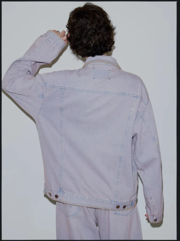 Person seen from behind in a pale lavender denim jacket and matching jeans, raising one hand against a gray backdrop