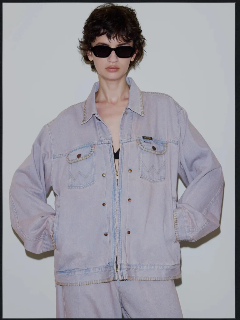 Model wearing black sunglasses and a pale lavender denim jacket with flap pockets, standing hands on hips against a gray