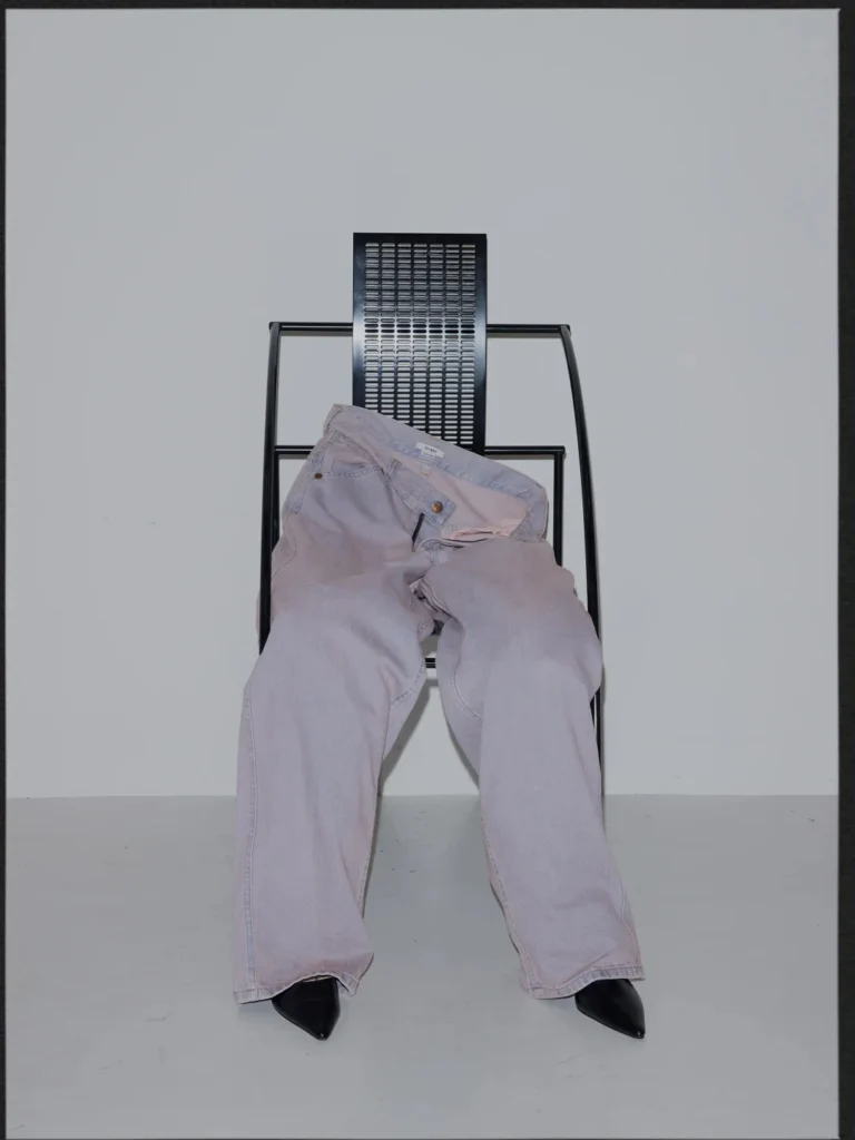 Pale pink wide-leg jeans draped over a black metal chair, with black pointed shoes beneath them in a bare studio setup