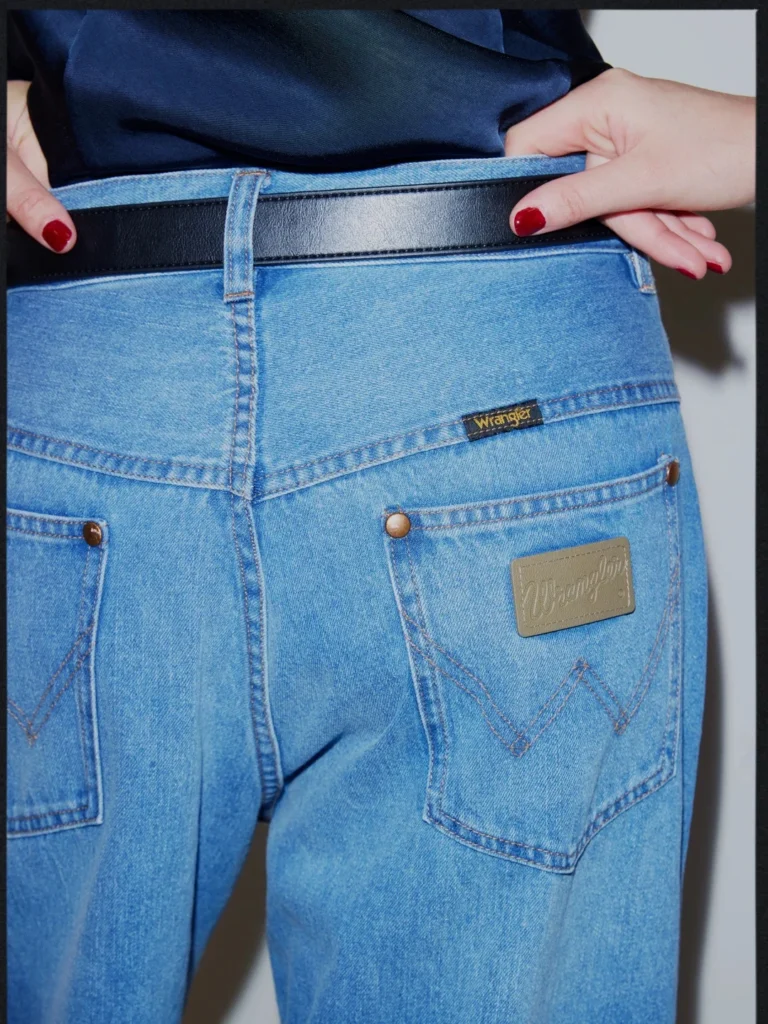 Close-up of light-blue Wrangler jeans with black belt, back pockets, stitched details, and hands with red-painted nails