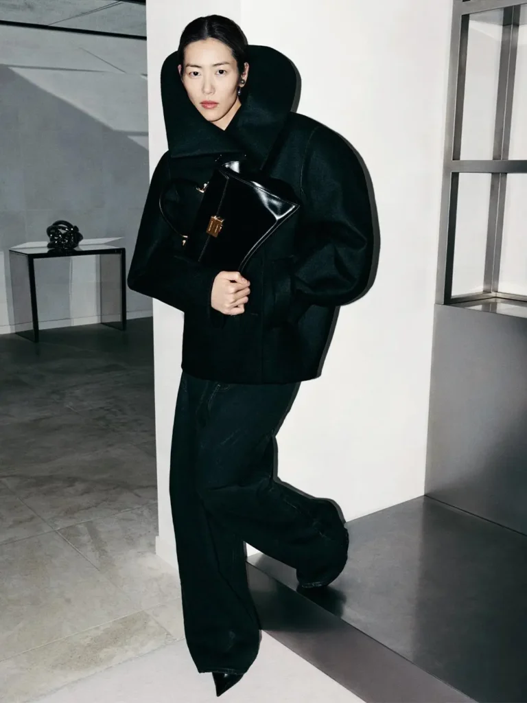 Person in a black oversized coat and wide-leg trousers holds a glossy black clutch in a modern interior