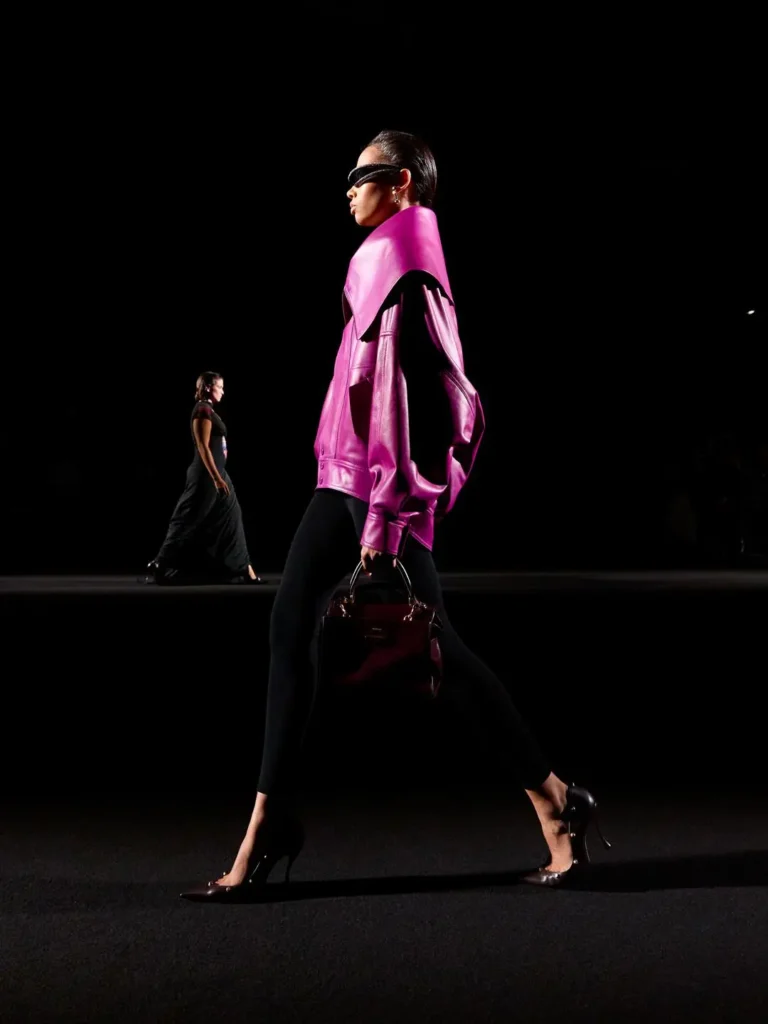 Model strides in a vivid magenta leather jacket, black leggings, sunglasses and heels, carrying a dark handbag on a dim