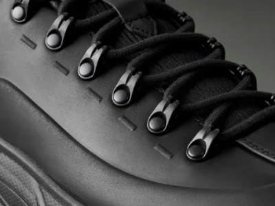 Close-up of a black shoe with thick laces, metal eyelets, leather panels and a textured rubber sole against a dark background
