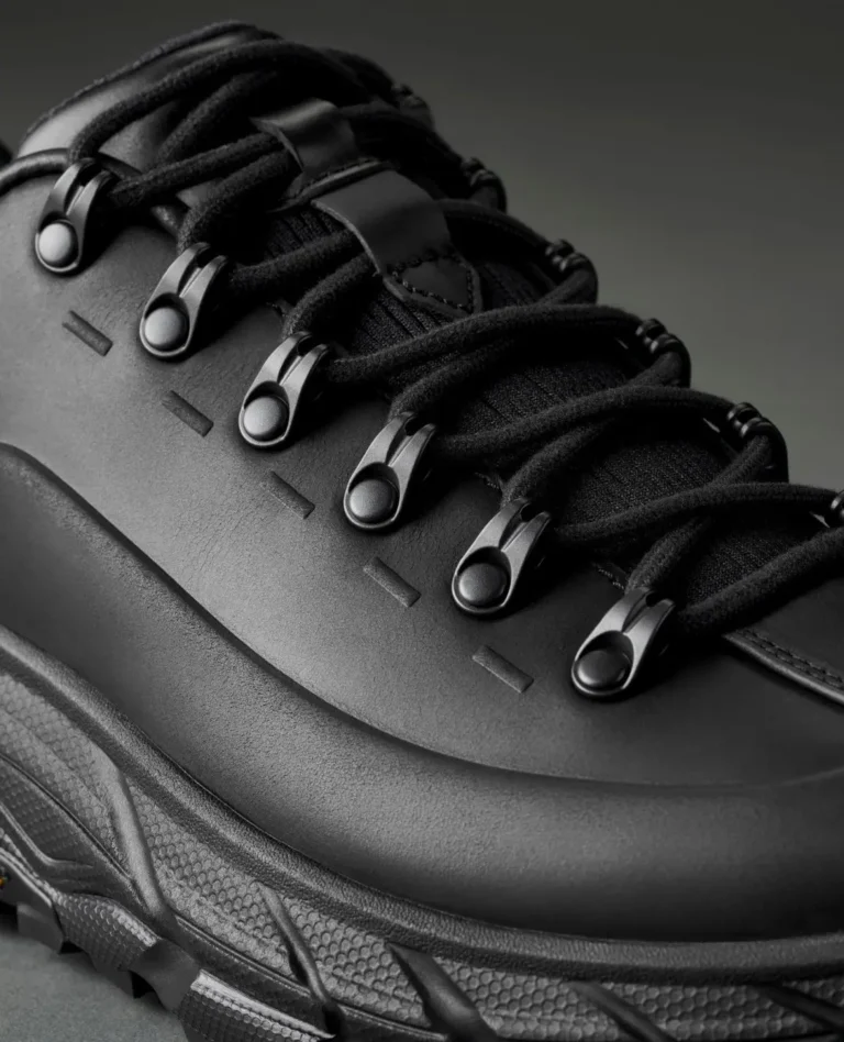 Close-up of a black shoe with thick laces, metal eyelets, leather panels and a textured rubber sole against a dark background
