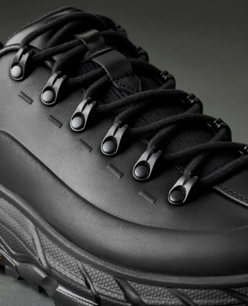 Close-up of a black shoe with thick laces, metal eyelets, leather panels and a textured rubber sole against a dark background