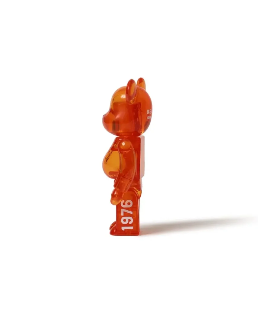 Translucent orange bear-shaped figurine standing in profile, with “1976” printed on its leg, casting a long shade on white