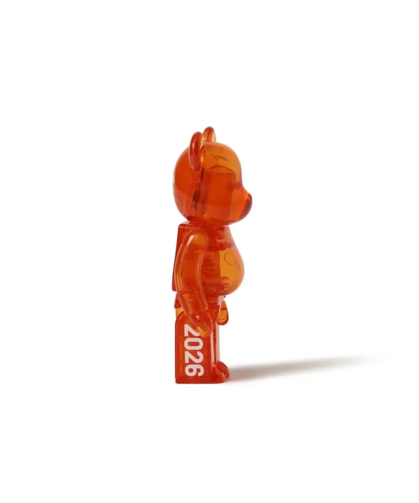 Translucent orange bear-shaped figurine standing in profile, marked “2026,” casting a long shade on white background
