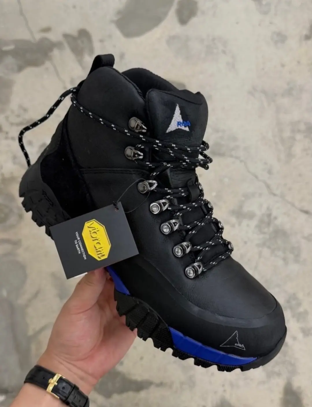 A hand holds a black high-top hiking boot with blue sole accents, metal eyelets, patterned laces, and a hangtag