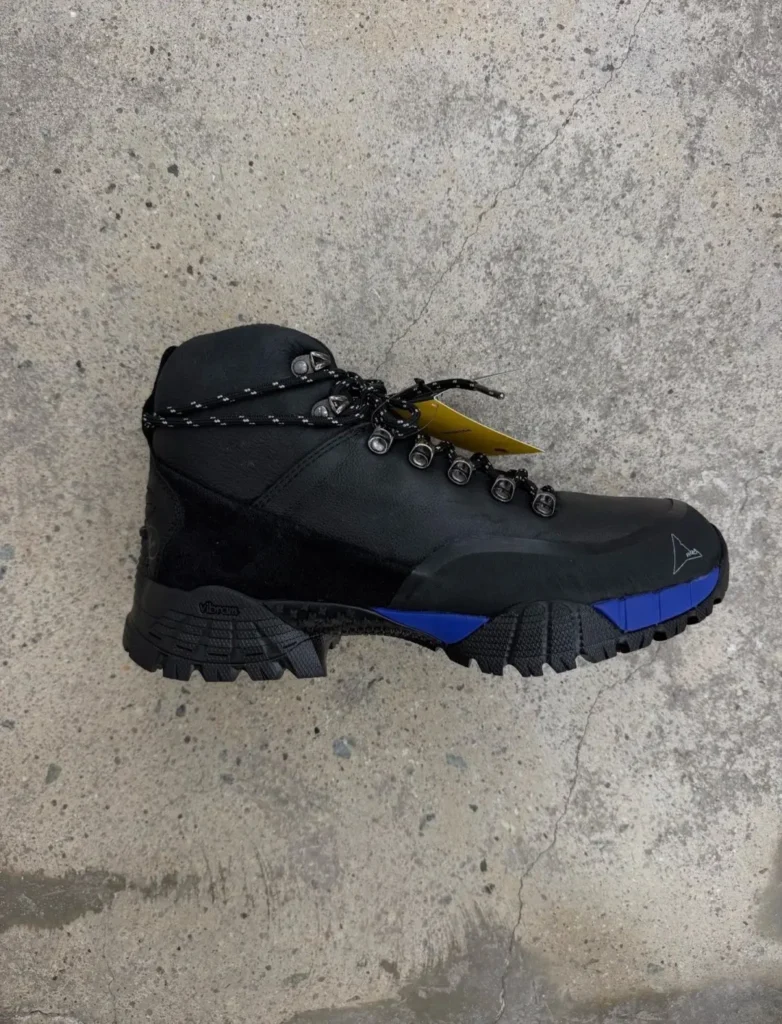 Black hiking boot with blue sole accents, metal eyelets and laces, photographed on a concrete floor