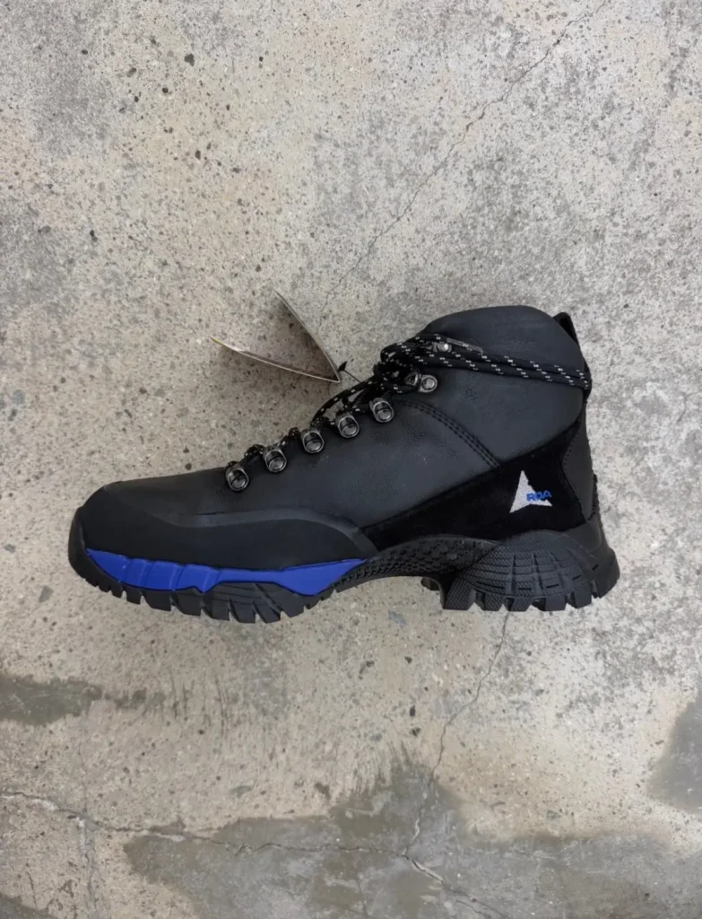 Black leather hiking boot with blue sole accents, metal eyelets and a small blue-and-white side logo on concrete surface