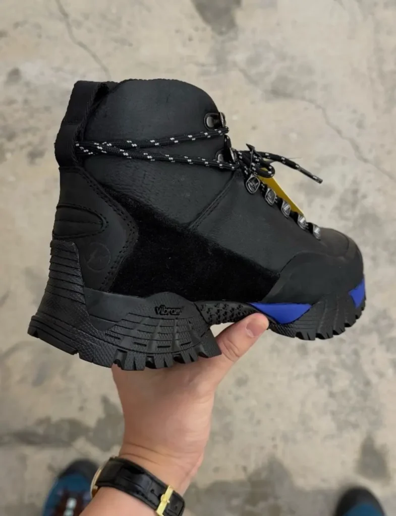 Black high-top hiking boot with rope laces, yellow accents and blue sole, held above a concrete floor