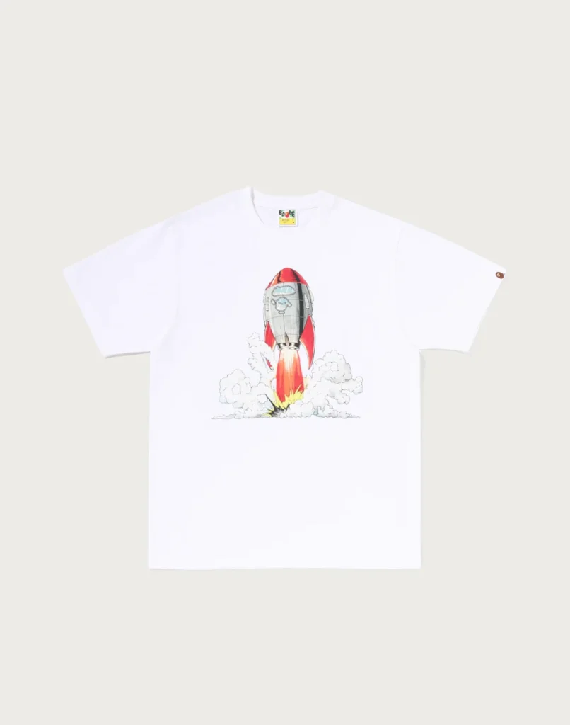 White T-shirt featuring a colorful rocket launching through clouds, laid flat against a pale background