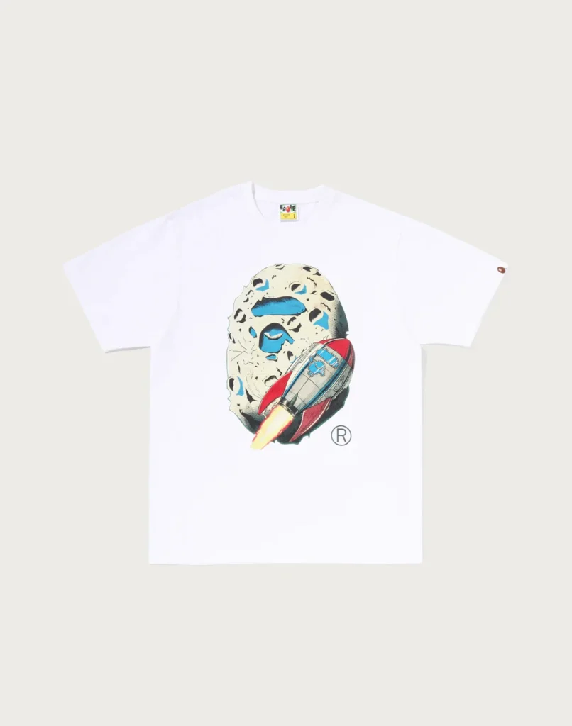 White T-shirt with a cartoon speckled face graphic and colorful rocket, laid flat against a pale background