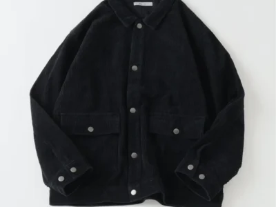 Black corduroy jacket with pointed collar, silver buttons, flap pockets and cuff tabs, laid flat on white background
