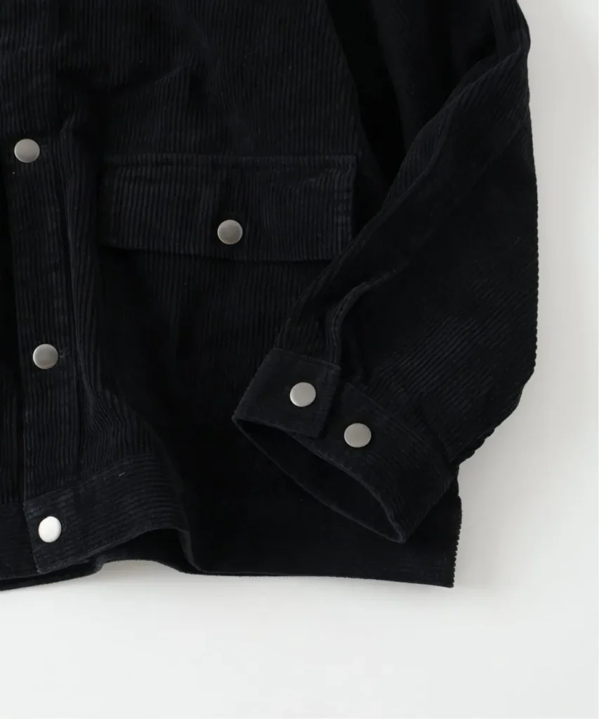 Close-up of a black corduroy jacket with flap pocket, silver buttons, and rolled sleeves against a white background