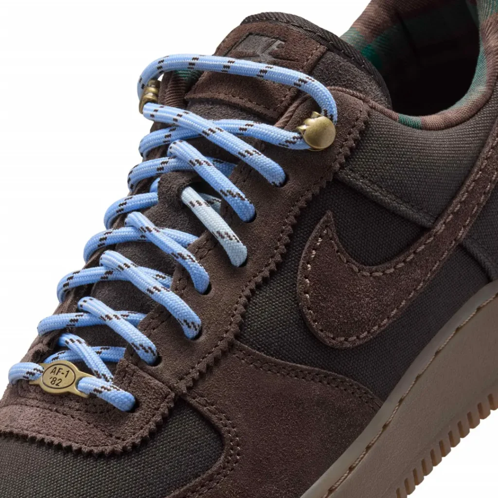 Close-up of a brown and black sneaker with light blue patterned laces, brass lace hardware, and a gum sole on a white