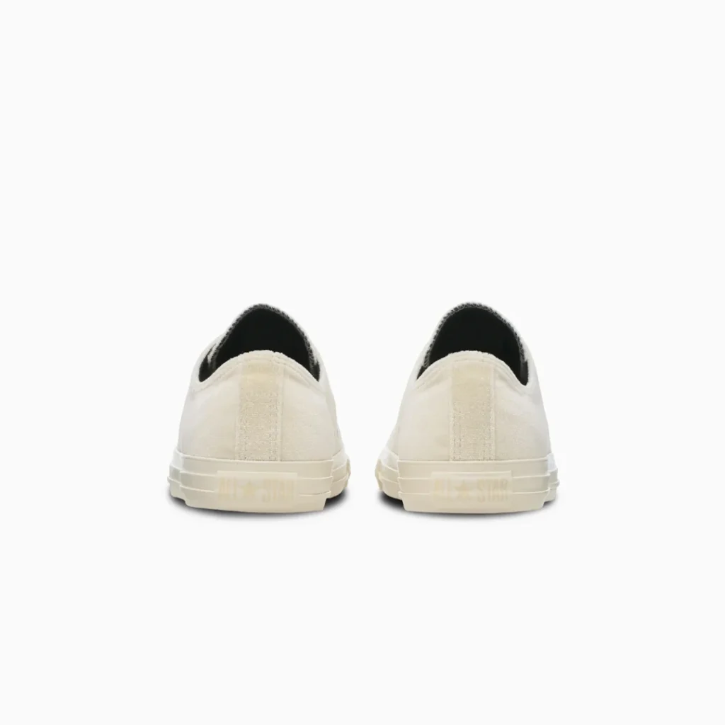 Rear view of a pair of cream canvas sneakers with black interiors and pale rubber soles against a white background