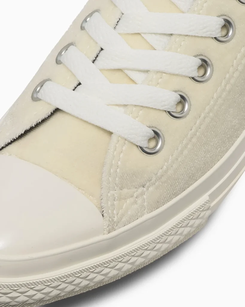 Close-up of a cream canvas sneaker with white laces, silver eyelets, and a textured rubber sole against a white background