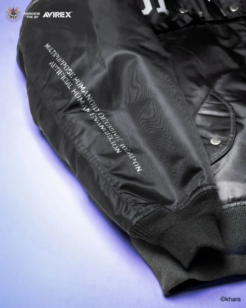 Close-up of a black MA-1 bomber jacket sleeve with white printed text, ribbed cuff, and snap pocket on a pale background