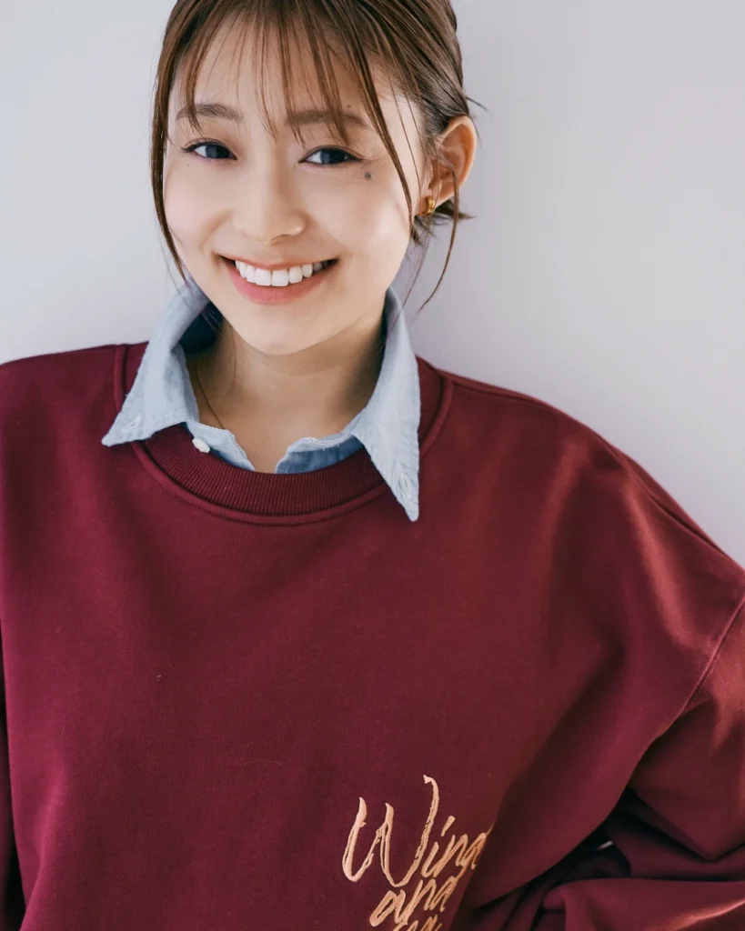 Smiling woman in a burgundy sweatshirt over a light blue collared shirt, posed against a plain wall