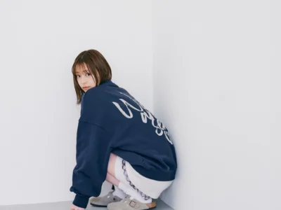 Person crouching in a white corner, wearing an oversized navy sweatshirt, white socks and gray buckle shoes