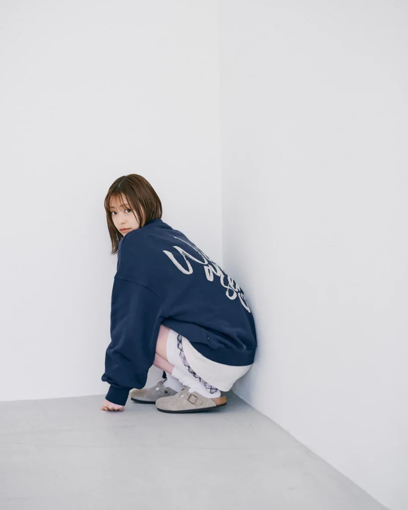 Person crouching in a white corner, wearing an oversized navy sweatshirt, white socks and gray buckle shoes