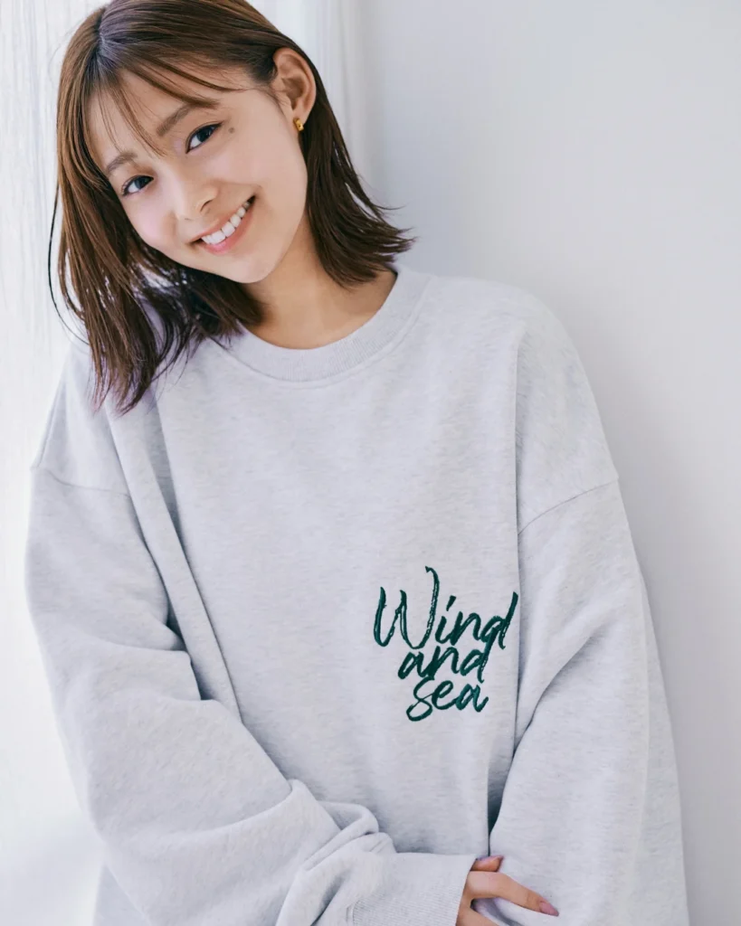 Smiling woman with shoulder-length brown hair wears a light gray sweatshirt reading “Wind and Sea” indoors against a white