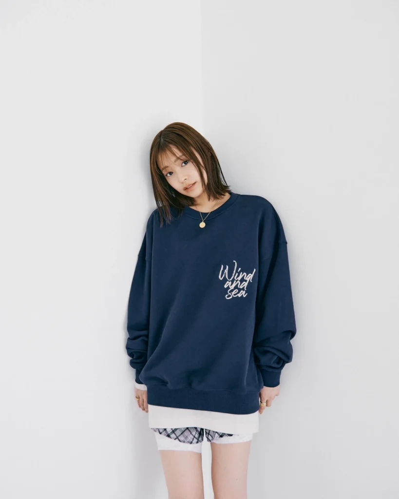 Woman leans against a white wall wearing an oversized navy sweatshirt with white “Wind and Sea” lettering and layered shorts