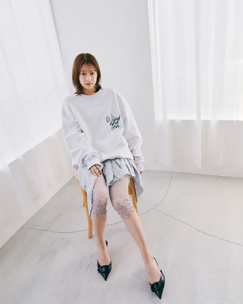 Woman seated in a bright room wearing a white sweatshirt, striped skirt, lace-patterned tights and black pointed flats