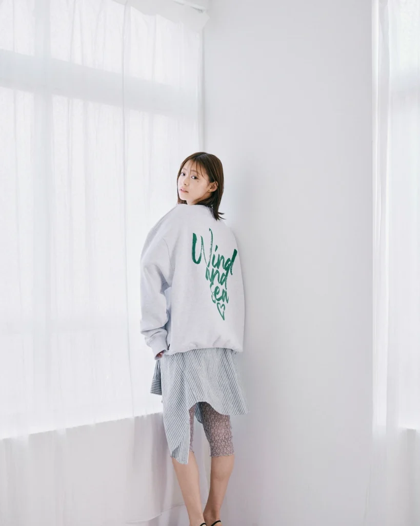 Woman looking over her shoulder in a light gray sweatshirt with green lettering and a striped asymmetrical skirt by sheer
