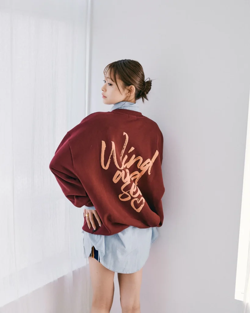 Person looking over her shoulder in a burgundy sweatshirt with peach script, layered over a blue shirt and shorts indoors