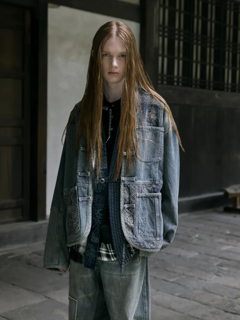 Person with long red hair wears a layered blue denim jacket with patterned panels and multiple pockets, standing indoors